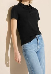 Svart polo shirt med krage, korta ärmar och tre knappslå, i kombination med ljusblå denimjeans. Slät tygtextur.