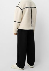 Fleecejacka i krämfärg med svart sömnad, fyrkantig design. I kombination med lösa svarta byxor och ljusfärgade sportskor.