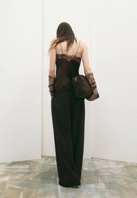 Mujer con cabello largo que lleva un camisón negro con encaje, pantalones de pierna ancha, guantes translúcidos y sostiene un bolso grande y texturizado, de pie frente a un fondo blanco.