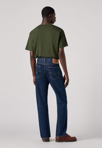 Hombre con camiseta de color verde oscuro, jeans rectos azules con parche de cuero marrón y zapatos marrones, de pie mirando hacia atrás sobre un fondo liso.