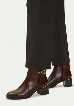 Stivaletti in pelle marrone con tacco a blocco, zip laterale e dettaglio con fibbia, indossati con pantaloni su misura marrone scuro.