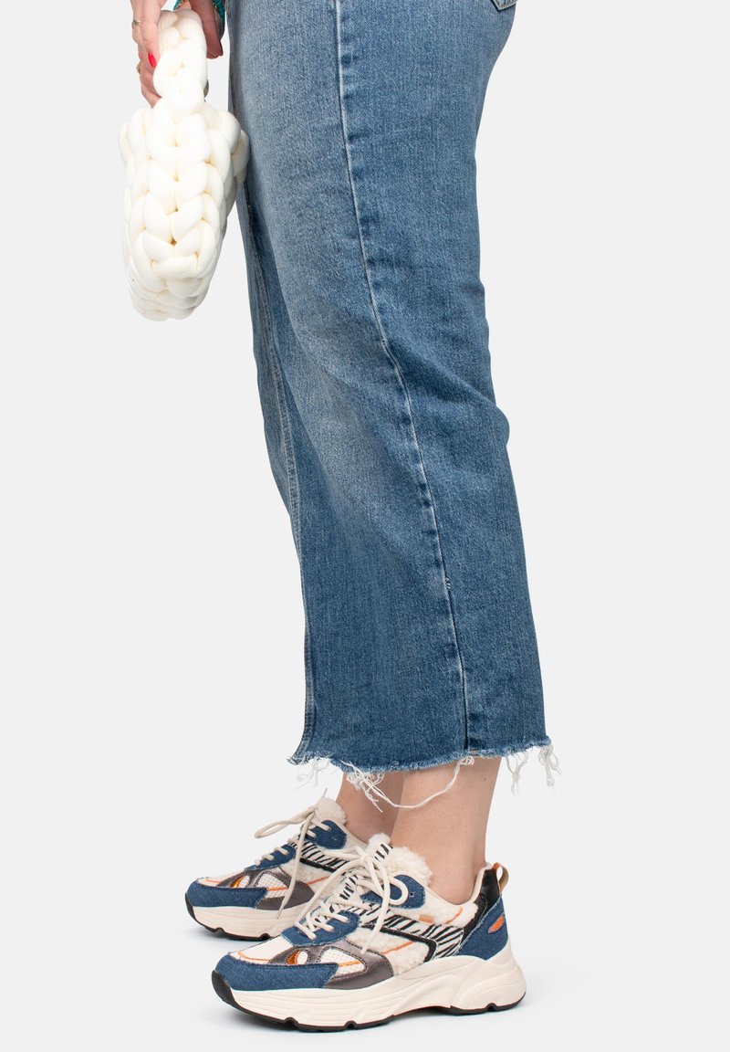 Les baskets présentent un mélange de matériaux avec un schéma de couleurs bleu et crème, une tige texturée et une semelle épaisse. Le bas des jeans est effiloché.