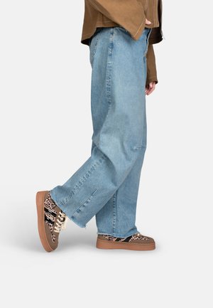 Personne portant un jean large bleu clair et des baskets marron à imprimé léopard avec des rayures noires, debout devant un fond uni.