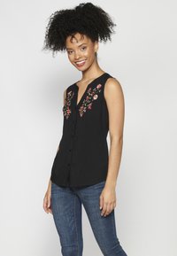 Blusa nera senza maniche con scollatura a V, caratterizzata da ricami floreali in tonalità di rosa e verde sul davanti, e bottoni al centro.