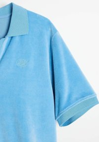 Jasnoniebieska koszulka polo wykonana z miękkiego materiału, z klasycznym kołnierzykiem i krótkimi rękawami. Haftowane logo w nieco ciemniejszym odcieniu niebieskiego.