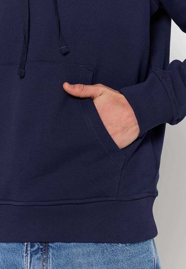 Sweat à capuche en coton bleu marine avec une poche avant, des poignets côtelés et un ourlet côtelé. Main partiellement insérée dans la poche. Texture lisse et unie.