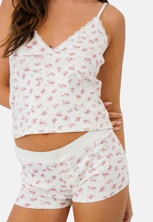 Femeie purtând un top de tip camisolă cu imprimeu floral alb și pantaloni scurți asortați cu nasturi, în picioare în fața unui fundal simplu și luminos.