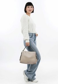 Beige leather bag with dual top handles, zip closure, and side pockets, carried by a person in a white knitted sweater and blue jeans.