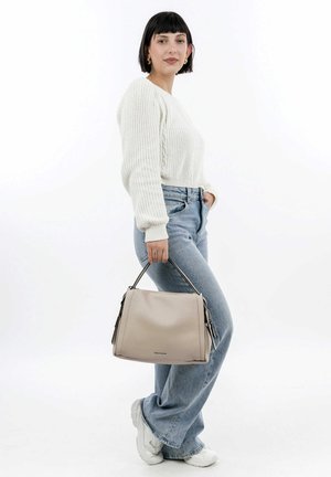 Sac en cuir beige avec deux poignées supérieures, fermeture éclair et poches latérales, porté par une personne en pull en tricot blanc et jeans bleus.
