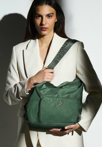 Borsa a tracolla verde con texture, dotata di un comparto principale con zip e tasca frontale. Presenta una tracolla larga con testo stampato. Superficie liscia, senza motivi.