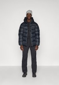 Veste matelassée noire avec texture quiltée, associée à un pantalon noir et un bonnet en tricot gris. Le mannequin se tient devant un fond neutre.