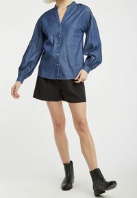 Jeansblus med en vertikal knappslå, långa puffärmar och en lätt v-ringning. Buren med svarta shorts och ankelstövlar.