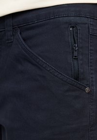 Navy fabric pants featuring a visible zippered pocket, smooth texture, and a button closure. Stitching lines and pocket shapes are prominent.