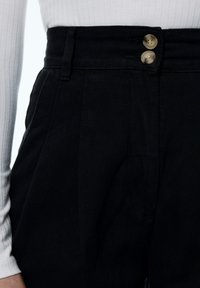 Zwarte high-waisted broek met twee grote knopen aan de voorkant, getailleerde pasvorm, opvallende zakken en een gestructureerde stofafwerking.
