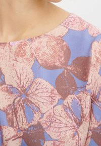 Zbliżenie na odzież z okrągłym dekoltem, ozdobioną dużym kwiatowym wzorem w odcieniach różu i brązu na jasnoniebieskim tle.