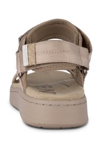 Beige sandals with textile straps, padded heel, and a flat sole. Features a textured footbed and a logo accent on the strap.