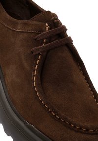 Scarpa in suede marrone con punta rotonda, caratterizzata da lacci marroni, cuciture color sabbia e una suola in gomma testurizzata. Superficie liscia e aspetto morbido.