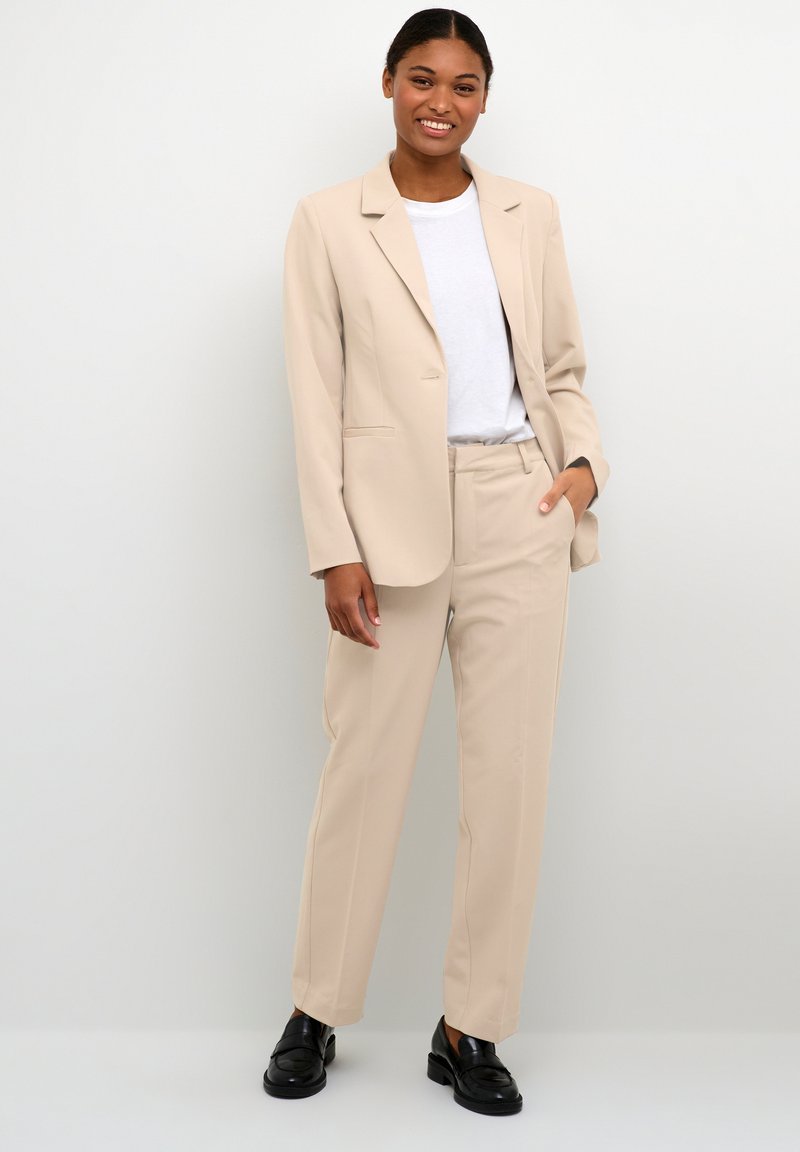Beige skräddarsydd kostym med en enknäppt blazer, raka byxor och en enkel vit t-shirt. Svarta loafers fulländar looken.