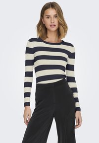 Gestreepte ribbed lange mouw top in crème en marineblauw, met een ronde hals en een getailleerde silhouette. Gecombineerd met zwarte wijde broek.