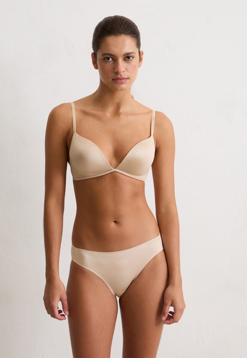 Femme se tenant contre un mur neutre, portant un soutien-gorge couleur chair et une culotte de style bikini assortie, faisant face avec une expression neutre.