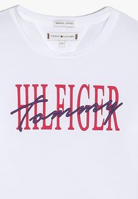 Biała koszulka T-shirt z organicznej bawełny z okrągłym dekoltem, na której widnieje napis "TOMMY" w granatowym kursywie oraz "HILFIGER" w pogrubionym czerwonym.