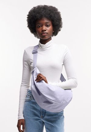 Sac bandoulière lilas en tissu doux, de forme incurvée et doté d'une sangle ajustable, tenu par une personne portant un col roulé blanc.