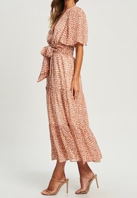 Maxime dress in textured, light pink fabric with a patterned design, V-neck, short sleeves, and a tied waist belt. Features a flowing silhouette.