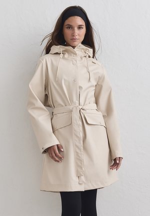 Giovane donna con lunghi capelli castani che indossa un impermeabile beige con cintura e cappuccio e leggings neri davanti a una parete chiara e uniforme.