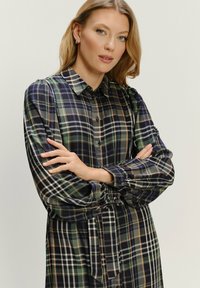 Žena s svetlými vlasmi oblečená v dlhé rukávy, gombíkovej, kárované šaty v zelených, modrých a béžových odtieňoch, stojí s prekríženými rukami.