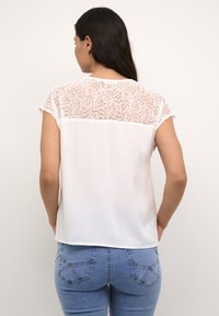 Blusa bianca con bordo in pizzo sulle spalle e sulla schiena, con maniche corte e un tessuto liscio e morbido. Abbinata a jeans blu.