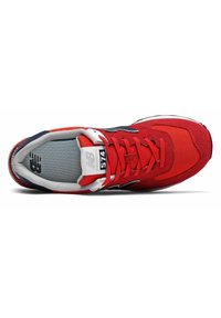 Zapatilla deportiva roja con materiales de ante y malla, que presenta un cuello acolchado, cordones blancos y un logo negro en el costado. Diseño clásico.