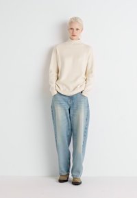 Cremfarbener Rollkragenpullover kombiniert mit hellblauen, locker sitzenden Jeans mit ausgeblichenen Details, getragen mit beigen Stiefeln.