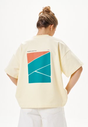 Kobieta z upiętymi włosami nosząca kremową, oversize koszulkę z geometrycznym wzorem kortu tenisowego w kolorach teal i pomarańczowym na plecach.