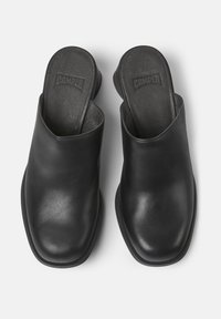 Een paar zwarte leren instapschoenen met vierkante neuzen en lage hakken, met ingegraveerde merkinleggers met de tekst "CAMPER."