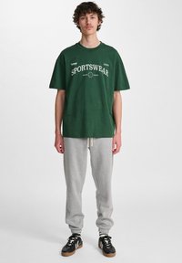 Zielona bawełniana koszulka z białym napisem "SPORTSWEAR", zestawiona z szarymi spodniami dresowymi i czarnymi sneakersami z brązowymi akcentami. Prosty, luźny krój.