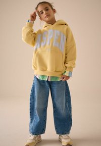 Żółta bluza z kapturem z nadrukiem "ASPEN", noszona na paskowanym t-shircie, w połączeniu z luźnymi niebieskimi dżinsami i beżowymi sneakerami.