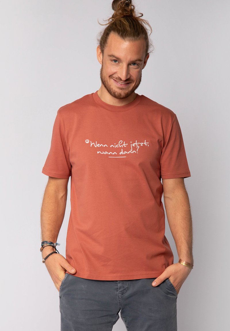 Homme avec un chignon portant un T-shirt couleur rouille avec le texte allemand « Wenn nicht jetzt, wann dann ? » et un pantalon gris, les mains dans les poches.