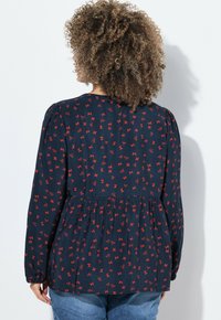 Marinebluse mit langen Ärmeln und gerafftem Bund, mit einem roten Schleifenmuster und subtiler Textur, getragen mit blauen Jeans.