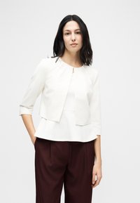 Femme aux cheveux foncés portant une veste blanche courte par-dessus un haut blanc et un pantalon marron foncé, debout avec une main dans la poche.