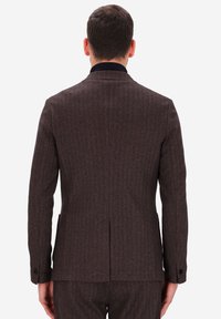 Uomo che indossa una giacca di tailleur marrone a spina di pesce ben aderente e pantaloni coordinati, in piedi di spalle su uno sfondo bianco uniforme.