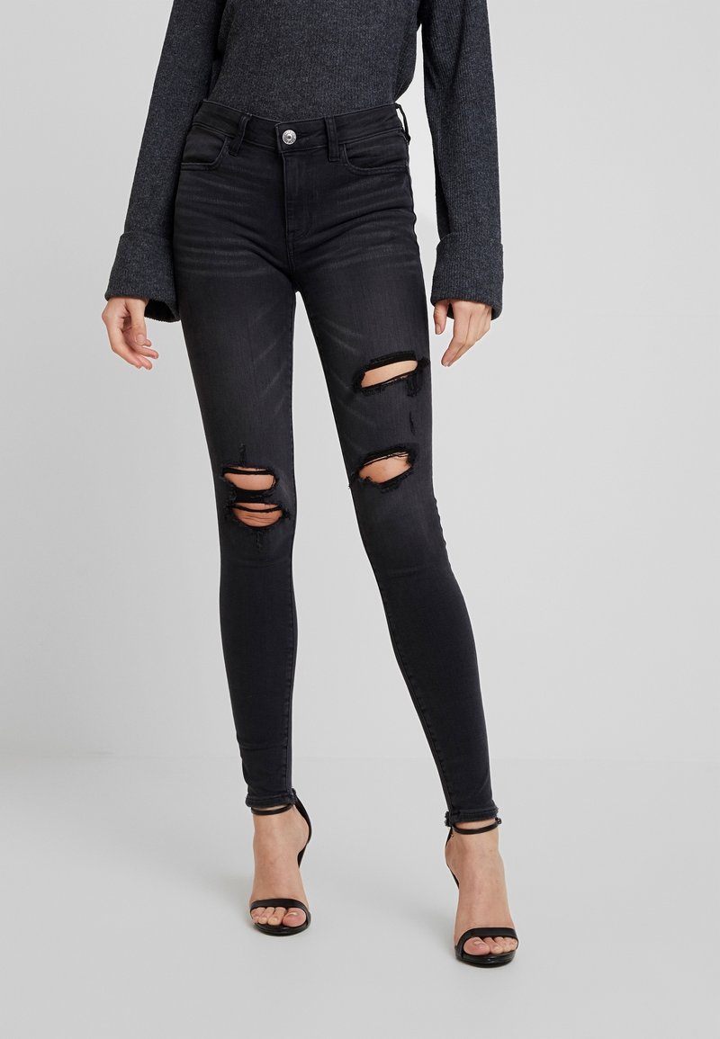 Kvinde iført sorte skinny jeans med høj talje og tre vandrette revner på forsiden af lårene, kombineret med en mørkegrå langærmet top og sorte åbne hæle.