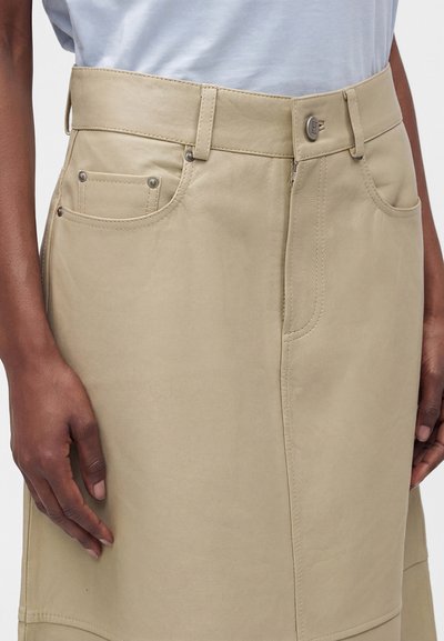 Gros plan d'une personne portant une jupe en cuir beige avec des poches avant et une ceinture boutonnée, associée à un haut blanc.
