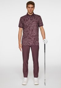 Mörkröd golfpolo med vitt konturmönster, korta ärmar och krage; matchad med mörkröda byxor och vita sneakers, och håller en golfklubba.