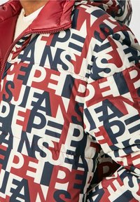 Puffjacka med ett djärvt grafiskt mönster i röd, marinblå och crème. Materialet är glänsande; designen inkluderar en huva och dragkedjestängning.