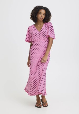 Göndör hajú nő, aki rózsaszín, fekete pöttyös, bokáig érő ruhát és fekete szandált visel, egyszínű világos háttér előtt áll.