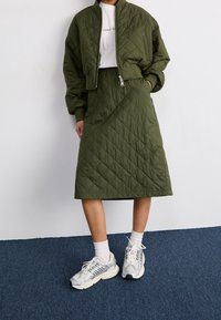 Olivově zelená prošívaná bomber bunda, odpovídající prošívaná midi sukně a stříbrné sportovní boty. Bílé tričko s grafikou pod tím, s žebrovanými manžetami.