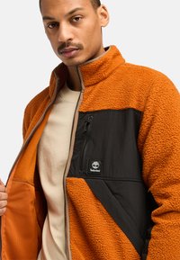 Orange Fleecejacke mit schwarzen Nylonakzenten, ausgestattet mit einer Reißverschlusstasche und einem Logo. Das Design beinhaltet einen strukturierten, warmen Stoff.