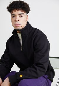 Sort halv-zip pullover sweatshirt med en blød tekstur, der har et gult logo på manchetten. Modellen sidder foran en lys baggrund.