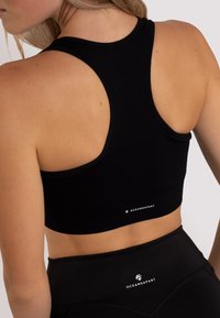 Czarny stanik sportowy z wycięciem na plecach, prążkowaną fakturą i detalem logo na dole. Materiał wydaje się elastyczny i dopasowany.