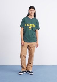 Zielona bawełniana koszulka z żółtym logo "Champion", w połączeniu z beżowymi spodniami i czarnymi sneakersami z białymi podeszwami i sznurowadłami.